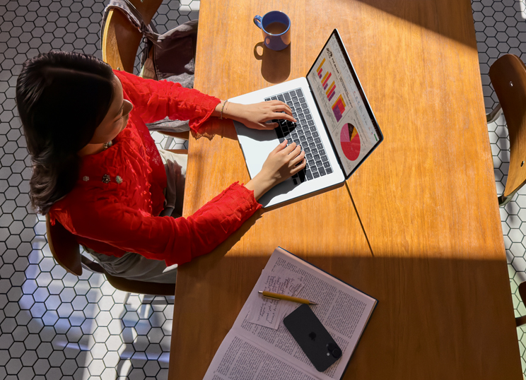 Une personne assise à une table utilise un MacBook. Une tasse, un téléphone intelligent et des papiers sont placés à proximité.