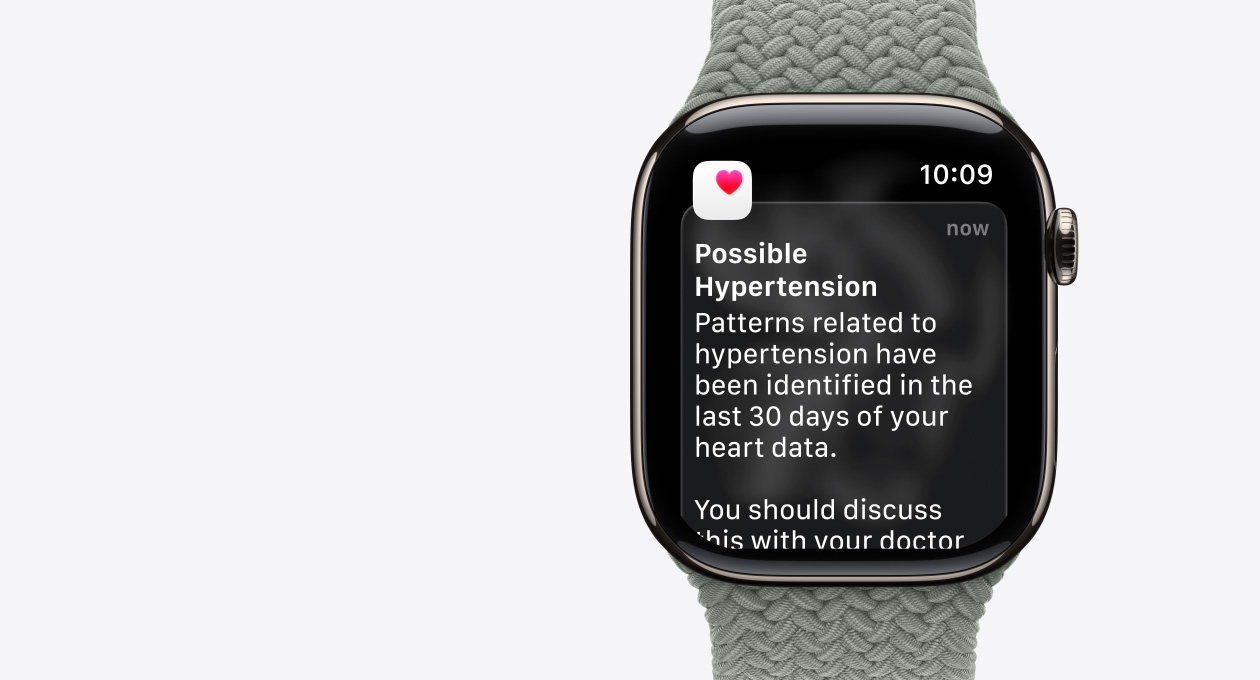 Apple Watch Series 11，原色鈦金屬錶殼，螢幕顯示可能高血壓通知，以及白色盒子內有粉紅色心心的圖示；右側外觀，數碼錶冠，配搭灰綠色編織單圈手環錶帶