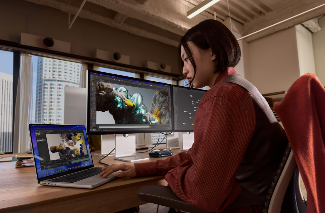 一個人在辦公桌前工作，MacBook Pro 連接外接顯示器，兩個螢幕皆顯示 3D 算圖畫面，由後方窗外可看見外面的建築物
