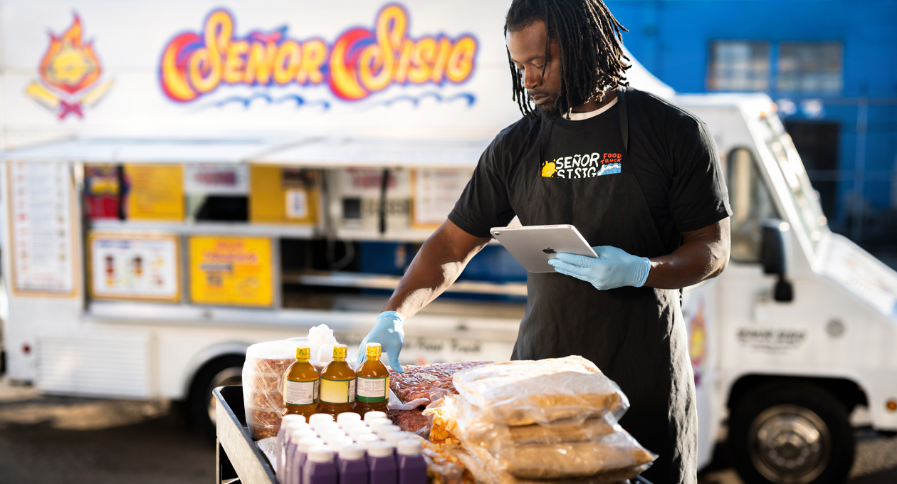 A person in a branded t-shirt and gloves reviews an iPad while prepping food at a table, a food truck in the background
