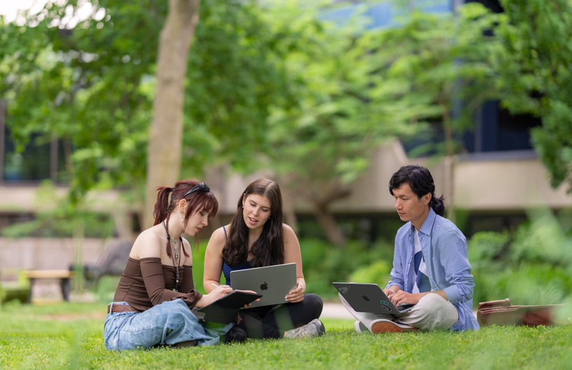 Three students sit in the grass on an outdoor campus, one using a MacBook while another looks on.