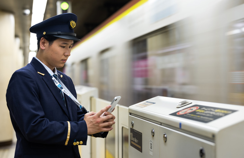駅係員がホームに立ち、動く電車の横でiPhoneでQRコードを読み取っている
