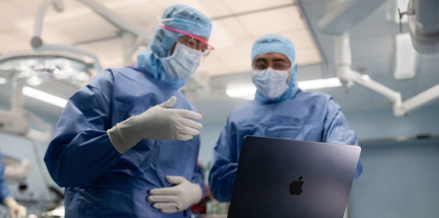 手術室でMacBookを見ている2人の外科医。1人が手でジェスチャーし、もう1人はそれを見ている