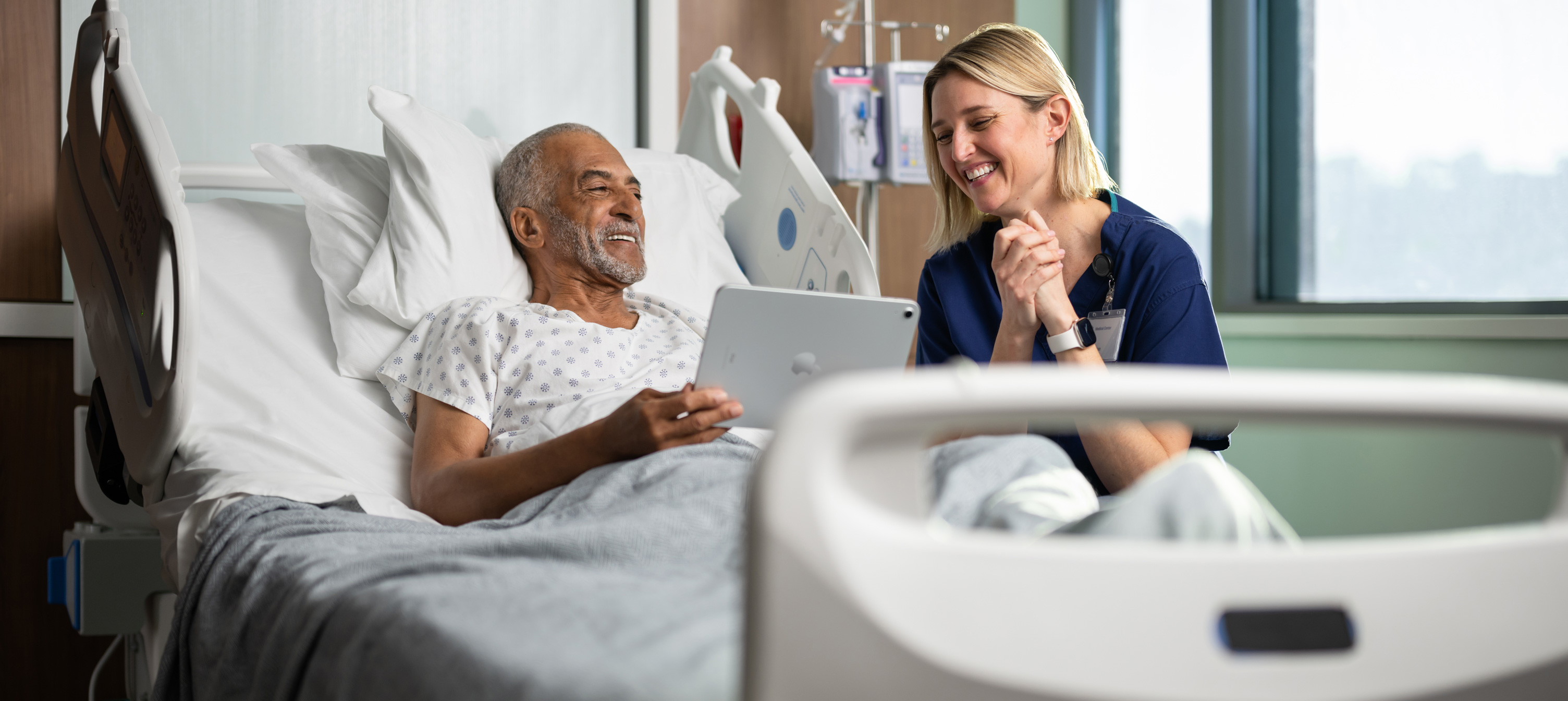 Un paziente in un letto d’ospedale. Sorride mentre guarda lo schermo di un iPad insieme a un’infermiera.