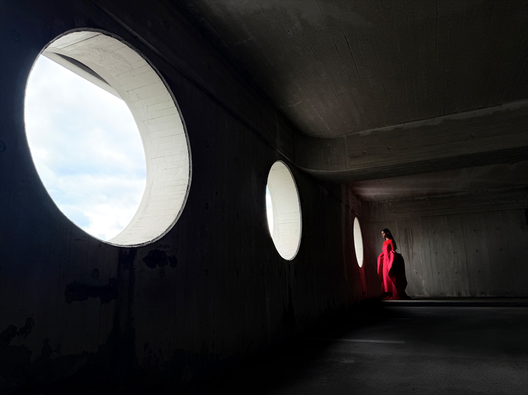 Une femme vêtue d’une longue robe rouge se fait photographier dans une pièce vide. L’app Appareil photo change de distance focale pour illustrer les capacités du système caméra pro, mettant en valeur: 24 mm