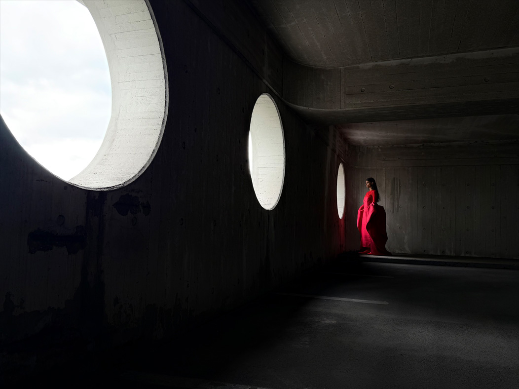 Une femme vêtue d’une longue robe rouge se fait photographier dans une pièce vide. L’app Appareil photo change de distance focale pour illustrer les capacités du système caméra pro, mettant en valeur: 28 mm